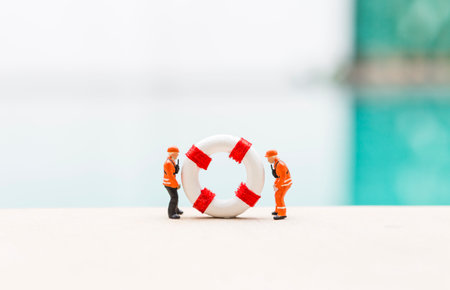 Two miniature worker in safety suite with lifebuoy over blurred swimming pool background, safety industry, outdoor day lightの写真素材