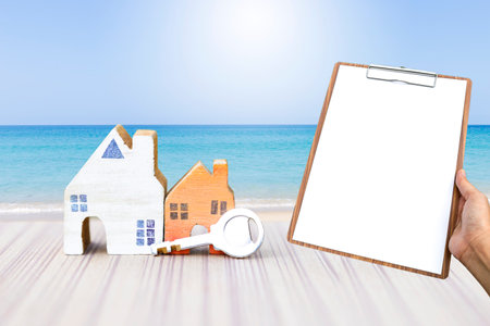 Miniature wooden house with key and blank clipboard over tropical beach background, property report, real estate business and investmentの写真素材