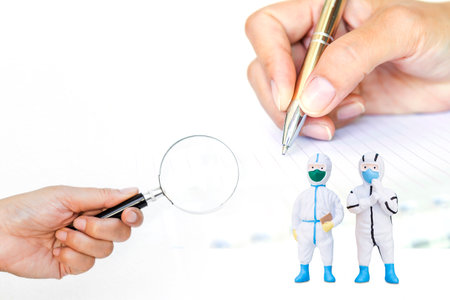 Miniature doctor wearing protection suit with magnify glass in girlhand over hand writing report background, health care and medical concept, doctor in PPE suit to protect againstの写真素材