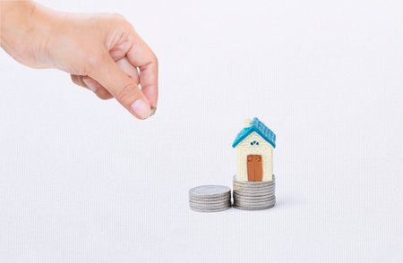 Miniature house on coin stack with hand holding coin on white texture background, property investment, real estate business, buy new houseの写真素材