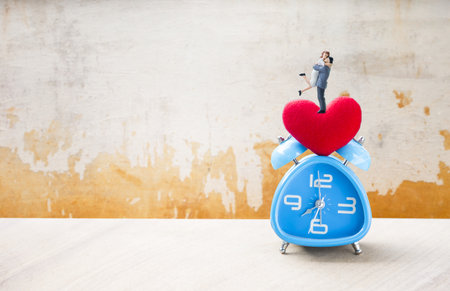 Miniature couple with red heart on blue alarm clock with space on old cement wall background, love and romance concept, valentine background ideaの写真素材