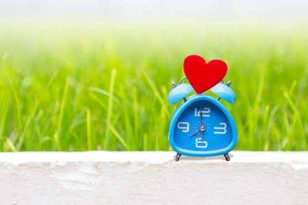 Red heart on blue alarm clock with space on blurred green garden background, time and love, valentine background ideaの写真素材