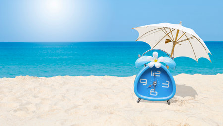Classic style blue alarm clock with plumeria flower and white beach umbrella with space on blurred tropical beach background, summer outdoor day light, holiday and vacation timeの写真素材