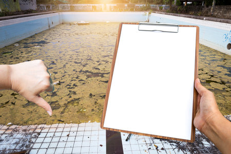 Blank report paper on clipboard in girl hand with thumbs down gesture over  old abandoned swimming...