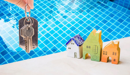 House key in girl hand with miniature house model over clear swimming pool water background, real...