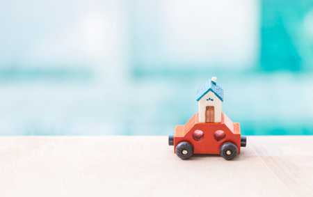 Miniature house model on red wooden toy car over blurred blue water background, property and real estate business idea