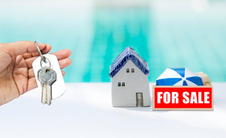 House key in girl hand with miniature house and for sale sign over blurred blue water background, buy beach house or summer house, real estate business conceptの写真素材