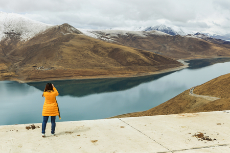Beautiful holy Yamdrok lake with snow mountain in background and a foreign tourist shooting a picture as foreground in a cloudy dayの写真素材