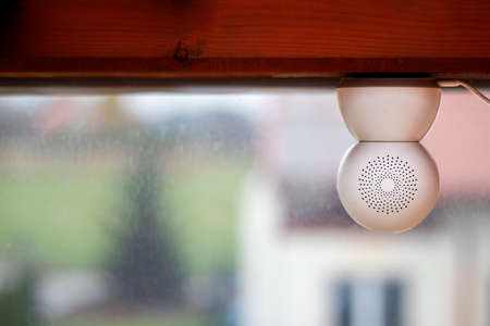 White CCTV camera mounted upside down on a wooden beam facing outside the window. The camera has a USB cable plugged into it and some patterned holes on it's back.の写真素材