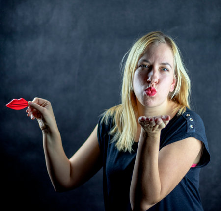 Caucasian blonde woman with red lipstick blowing a kiss while holding toy plastic lipsの写真素材