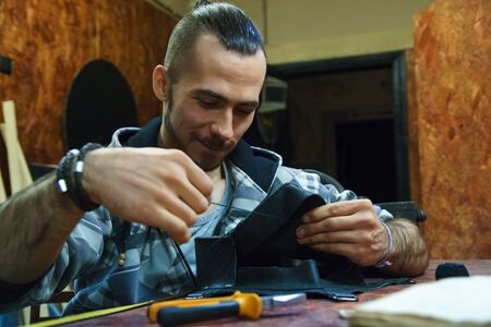 Tanner sews leather goods. Craftsman smiles while working. Making handmade bag. Male hands with...