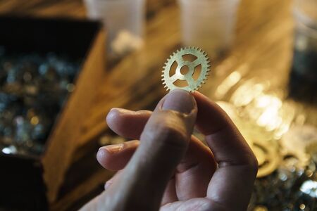 Watch repairer holds large gear. Big metal gearwheel on blurred workspace. Cogwheel on blurred...