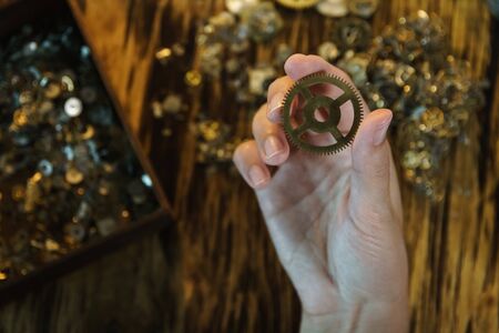 Watchmaker holds large gear. Cogwheel on blurred background of workplace. Pinion in female hand. Old cogwheel on wooden table background. Big metal gearwheel on blurred workspace.の写真素材