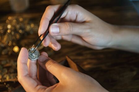 Female hands holding tweezers. Repairing vintage clocks. Hands with forceps shoot close up. Fingers with pincers and gears. Girl craftsman dismantles watch. Work at warm light of lamp.の写真素材