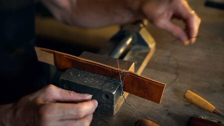 Man sews leather details clamped in vice. Craftsman works with needle and thread. Small business concept.の写真素材
