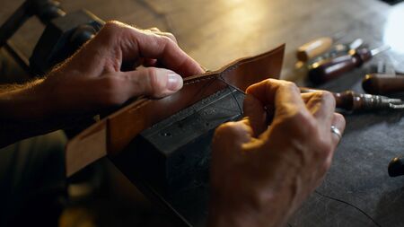 Man sews leather details clamped in vice. Craftsman works with needle and thread. Small business...