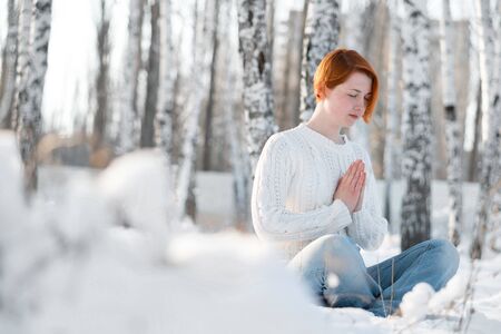 Young woman with hands clasped together in prayer. Lady with closed eyes sitting in winter snowy forest.の写真素材