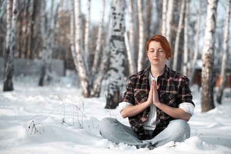 Girl praying to god in winter park. Woman sitting on snow in forest and meditating, copy space.の写真素材