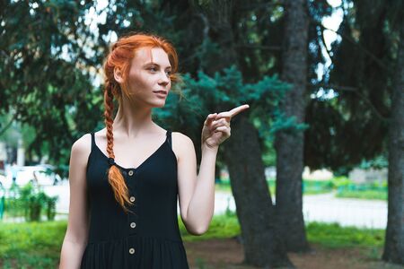 Ginger girl in black dress on nature background shows finger to side.の写真素材