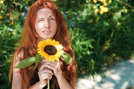 Cute ginger girl with sunflower look at sun in park. Spring mood.の写真素材
