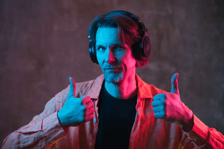 Attractive stylish man in big headphones colored by neon light show thumbs up on dark background. Modern music concept.の写真素材