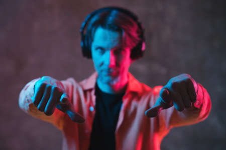 Adult hipster man in big headphones with red and blue neon lighting points to camera. Portrait with blurred face, soft selective focus. Modern music concept.の写真素材