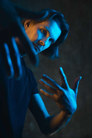 Adult stylish man in blue and yellow lighting looks at camera through hands. Neon light portrait.の写真素材