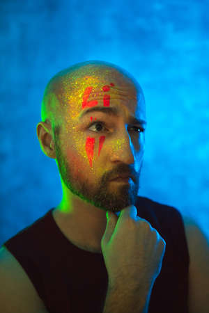 Pensive bald man with glowing fluorescent makeup on face in neon lighting. Ethnic, esoteric and art concept.の写真素材