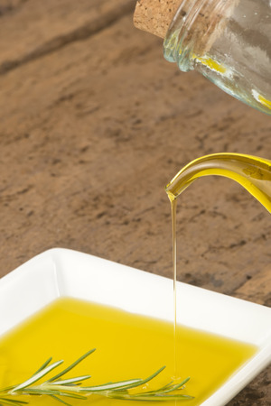 Glass jar pouring virgin olive oil in a plate with rosemary on rustic wooden tableの写真素材