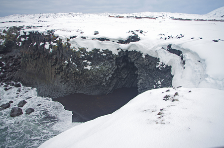 Icelandic winter landscape: Black volcanic rock under the snow at the seaside in front of snow-covered mountainsの写真素材