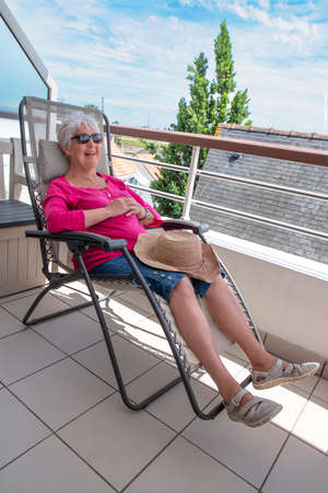 A smiling senior woman sitting in a deck chair on the terrace of a flatの写真素材