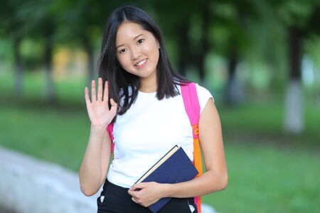 Girl female teenager with black hair in a bob white east Asian woman green park with a backpack background. Lady holds, dials, push your finger, smiling, looking at the camera and wavesの写真素材