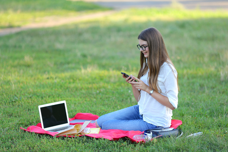 girl listening to music on the nature of the glasses. in the park for a laptop unravels headphones and holding a mobile phoneの写真素材