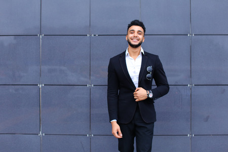 Young handsome businessman Arab Muslims is in the business center in black elegant dress on building background. Businessman man male looking at time walking goes and smiling.の写真素材
