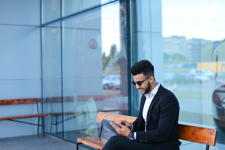 Entrepreneur smiling man  puts on sunglasses uses tablet and looks at. Young handsome businessman arab muslims in business center wearing dressed in black elegant suit on building background.の写真素材