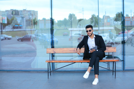 Entrepreneur smiling man  puts on sunglasses uses tablet and looks at. Young handsome businessman arab muslims in business center wearing dressed in black elegant suit on building background.の写真素材