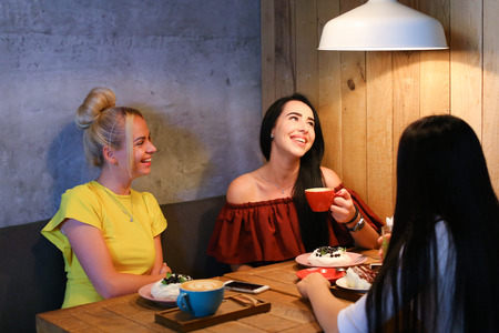 Concept female friendship, meeting, lunch. Three beautiful women sitting in a cafe smiling, talking, telling secrets, eat, drink coffeeの写真素材