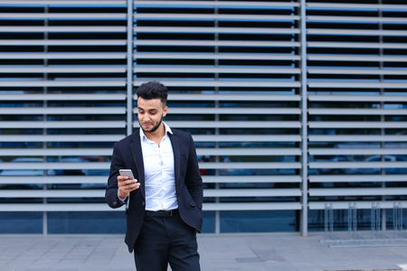 Entrepreneur uses phone mobile smart looking at the camera. Young handsome businessman arab muslims in business center wearing dressed in black elegant suit on building background.の写真素材