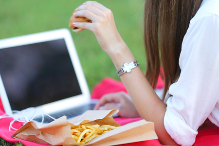 female in the park works behind laptop. beautiful girl with glasses eating fast food in nature and smiling.の写真素材