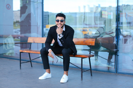 Entrepreneur smiling man  puts on sunglasses uses tablet and looks at. Young handsome businessman arab muslims in business center wearing dressed in black elegant suit on building background.の写真素材