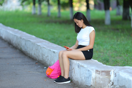 Girl female teenager with black hair in a bob white east Asian woman green park with a backpack background. Lady holds, dials, push your finger, smiling, looking into the camera, talking on the phone happy.の写真素材