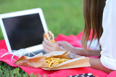 female in the park works behind laptop. beautiful girl with glasses eating fast food in nature and smiling.の写真素材