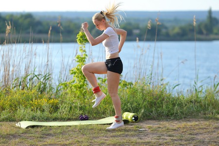 Young beautiful woman running shows press stomach workout training wearing top. Blonde female long hair weighted legs exercises engaged in sports be fit, strong form green background nature park.の写真素材