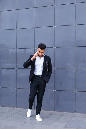 Entrepreneur man male looking at camera and smiling talks on mobile smart phone. Young handsome businessman arab muslims in business center wearing dressed in black elegant suit on building background.の写真素材