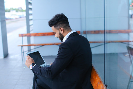 Entrepreneur uses laptop wearing sunglasses. Young handsome businessman arab muslims in business center wearing dressed in black elegant suit on building background.の写真素材