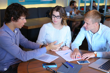 Two smart young business men one woman in suits sitting on table with documents, graphics, technology, laptop, phone, tablet. Team entrepreneurs negotiate, discuss issues, solve problems, agree, talkの写真素材