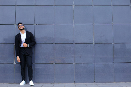 Young handsome businessman Arab Muslims is in the business center in black elegant dress on building background. Businessman man male looking at time walking goes and smiling.の写真素材