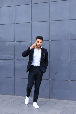 Entrepreneur man male looking at camera and smiling talks on mobile smart phone. Young handsome businessman arab muslims in business center wearing dressed in black elegant suit on building background.の写真素材