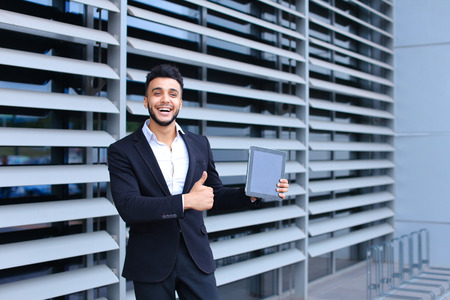 Young handsome businessman Arab Muslims is in the business center in black elegant dress on building background. Businessman man male looking waiting concept and smiling.の写真素材