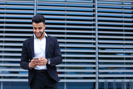 Entrepreneur uses phone mobile smart looking at the camera. Young handsome businessman arab muslims in business center wearing dressed in black elegant suit on building background.の写真素材